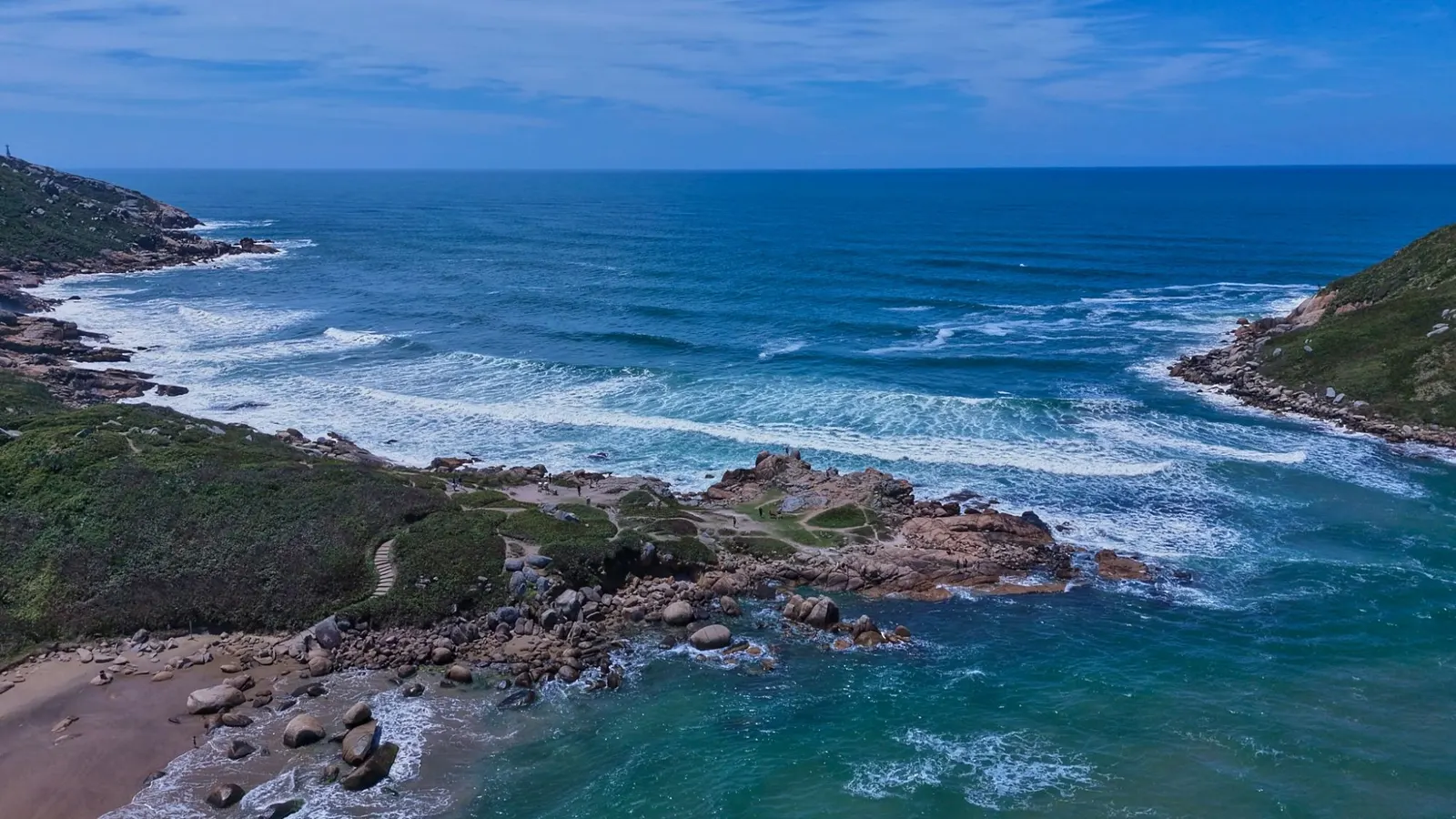 Vista aérea de uma enseada com costão rochoso e ondas chegando.
